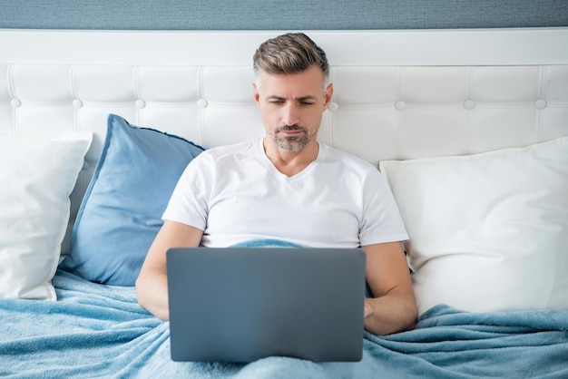 Homem maduro concentrado trabalhando no laptop na cama