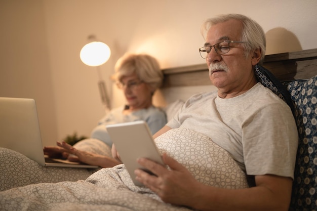 Homem maduro, deitado na cama e usando tablet digital enquanto navega na net no laptop