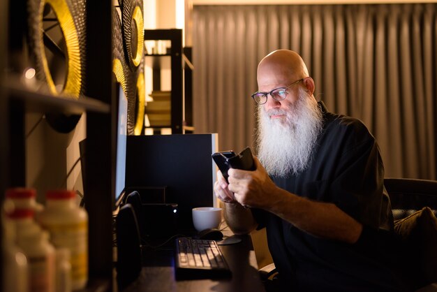 Homem maduro e careca barbudo usando o telefone enquanto trabalhava horas extras em casa tarde da noite
