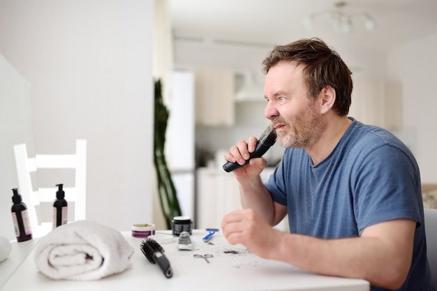 Homem maduro está raspando a barba com um barbeador elétrico em casa