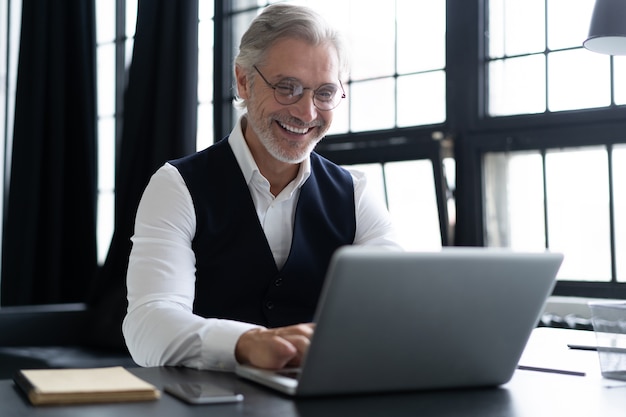 Homem maduro feliz com terno completo usando laptop enquanto trabalha em um escritório moderno