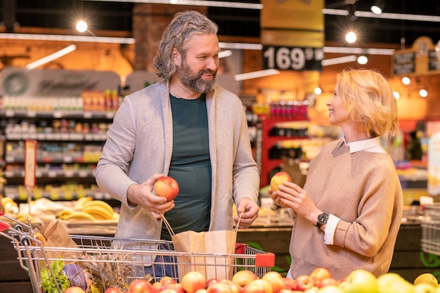 Homem maduro feliz e sua esposa escolhendo maçãs frescas enquanto estão em exposição com frutas no supermercado durante as compras