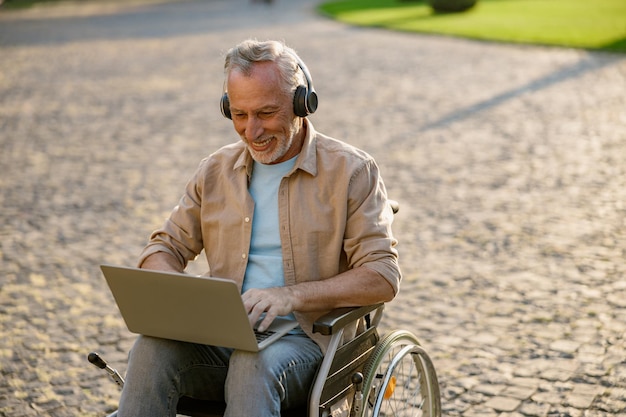 Homem maduro feliz recuperando paciente em cadeira de rodas usando fones de ouvido e trabalhando em um laptop ao ar livre