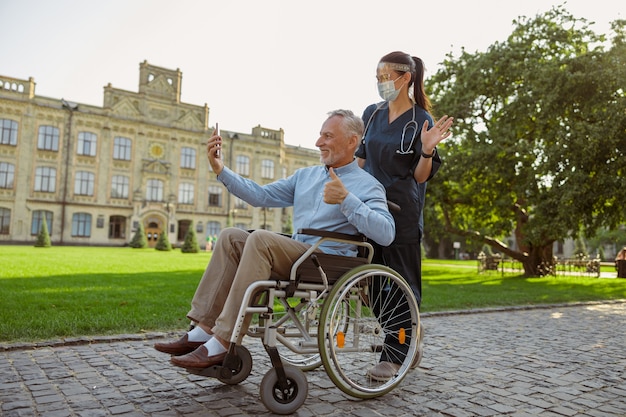 Homem maduro, recuperando paciente em cadeira de rodas, fazendo uma videochamada usando smartphone em uma caminhada com