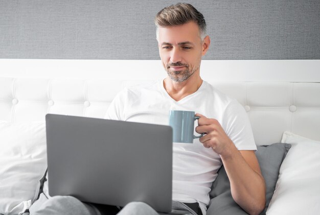 Homem maduro sorridente a usar computador na cama com café.