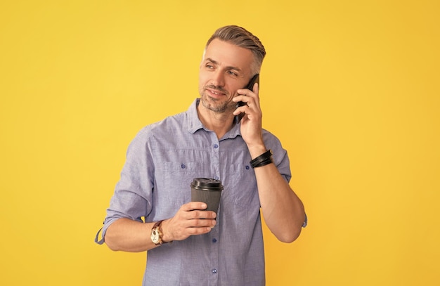 Homem maduro sorridente falando ao telefone com uma xícara de café na manhã de fundo amarelo