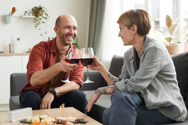 Homem maduro sorrindo para a mulher enquanto eles estão sentados no sofá e bebendo vinho em taças de vinho