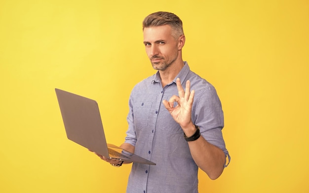 Homem maduro trabalhando on-line no laptop mostrando gesto ok em negócios ágeis de fundo amarelo