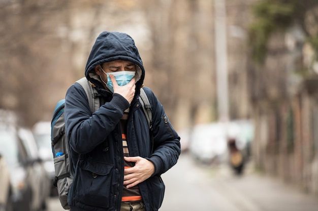 Homem maduro usa máscara facial e tosse em pé na cidade