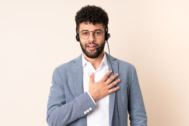 Homem marroquino de telemarketing trabalhando com um fone de ouvido isolado em uma parede bege surpreso e chocado ao olhar para a direita