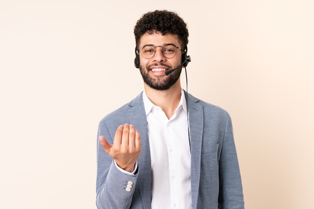 Homem marroquino do telemarketing trabalhando com um fone de ouvido isolado em bege, convidando para vir com a mão. Feliz que você veio