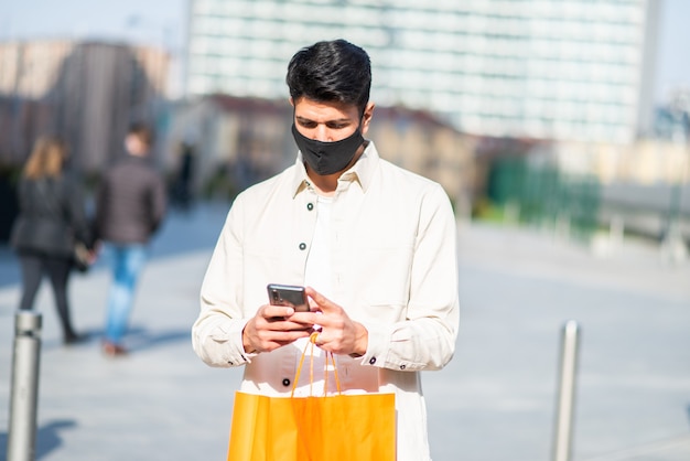 Homem mascarado carregando sacolas de compras enquanto usa um smartphone, conceito covid e coronavírus