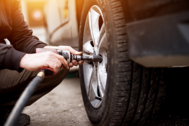 Homem mecânico de automóveis com chave de fenda elétrica, trocando o pneu do lado de fora. Serviço automotivo. As mãos substituem os pneus nas rodas. Conceito de instalação de pneus.