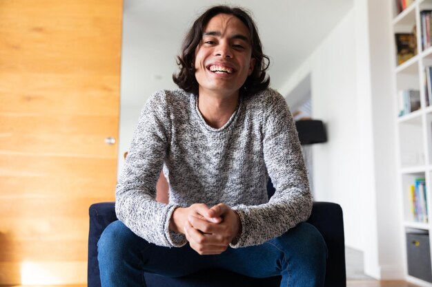 Foto homem mestiço feliz sentado no sofá, a fazer chamadas de vídeo e a sorrir, a passar tempo de qualidade sozinho em casa, a vida doméstica e o conceito de estilo de vida.