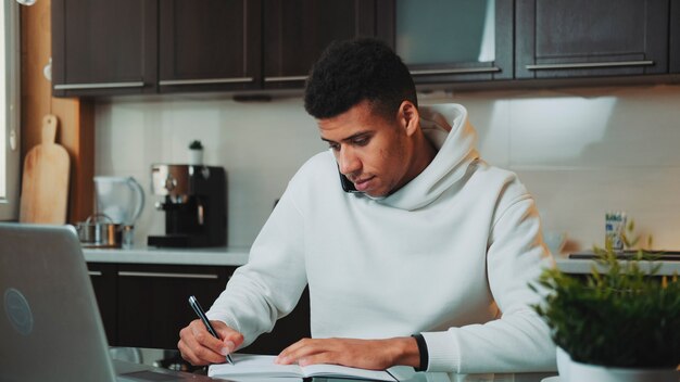 Homem misto falando pelo smartphone e trabalhando no computador na cozinha de casa