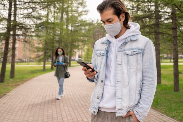 Foto homem moderno de jaqueta jeans e máscara facial em pé no parque e verificando o recado