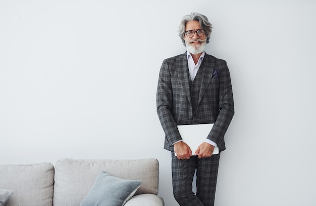 Foto homem moderno e elegante com cabelos grisalhos e barba dentro de casa de pé perto da parede com laptop nas mãos