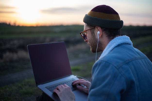 Homem moderno milenar trabalhando em um laptop com fones de ouvido no campo ao pôr do sol