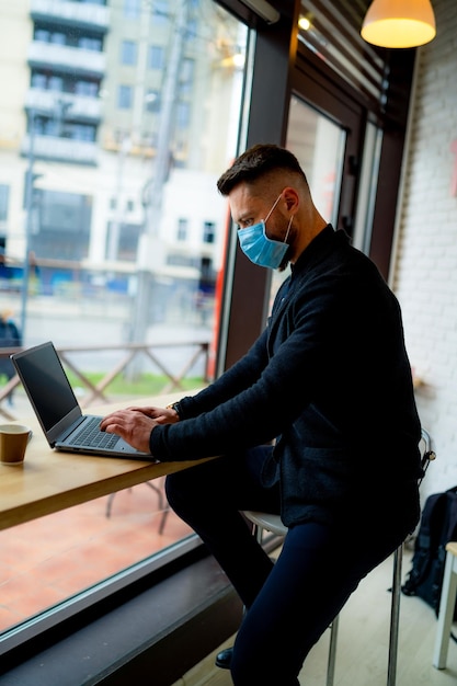 Homem moderno senta-se na cadeira do bar e trabalha no laptop em um café Gerente masculino usa máscara facial e digita no laptop pessoal
