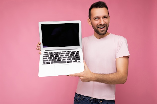 Homem moreno bonito e sorridente segurando um laptop olhando para a câmera em uma camiseta rosa isolada