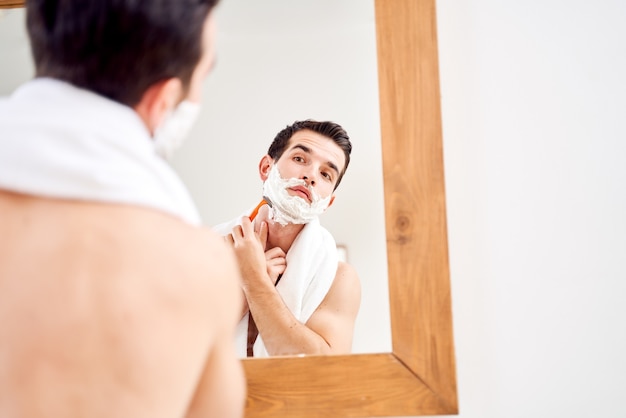 Homem moreno com espuma na barba faz a barba perto do espelho pela manhã