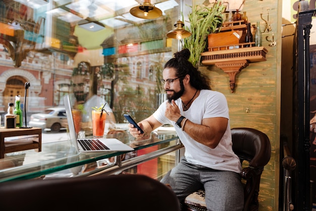 Homem moreno confiante usando seus gadgets enquanto está sentado em frente à janela do café