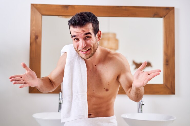 Foto homem moreno feliz com uma toalha no ombro em pé perto do espelho no banho matinal
