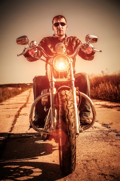 Homem motociclista vestindo uma jaqueta de couro e óculos de sol sentado em sua moto olhando o pôr do sol.