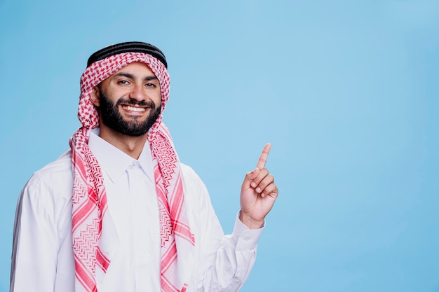 Homem muçulmano alegre vestido com manto tradicional apontando para cima com expressão sorridente. Produto de publicidade árabe despreocupado enquanto aparece na direção e olha para a câmera