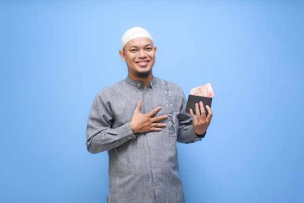 Foto homem muçulmano asiático segurando uma carteira cheia de dinheiro pronto para pagar zakat isolado em fundo azul