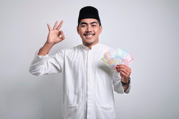 Foto homem muçulmano asiático sorridente em roupas brancas e boné de caveira preta sorrindo feliz enquanto segura o papel-moeda e mostra o gesto certo