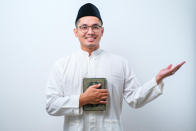Homem muçulmano asiático sorrindo segurando o livro al quran nas mãos e mostrando algo do seu lado sobre fundo branco