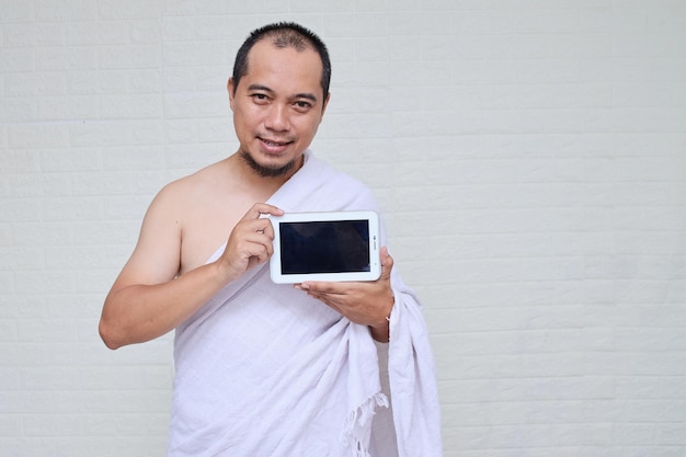 Homem muçulmano asiático vestindo roupas brancas de ihram e mostrando a tela do tablet