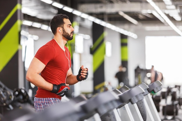 Homem musculoso barbudo usa camiseta vermelha faz exercícios de corrida cardio no ginásio