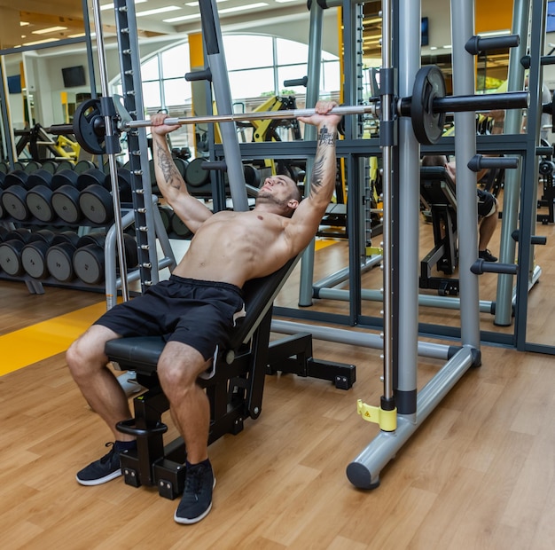 Homem musculoso com torso nu fazendo exercícios de supino com barra deitado em um banco inclinado na academia