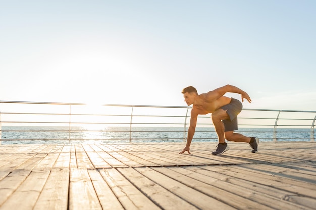 Homem musculoso com torso nu pronto para correr ao nascer do sol na praia