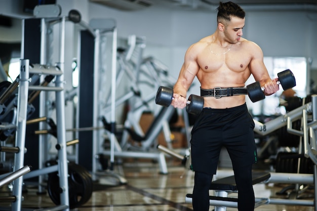 Homem musculoso e árabe treinando com ginásio moderno de halteres. Homens árabes de aptidão com torso nu, fazendo exercícios.