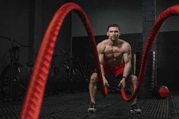 Homem musculoso fazendo exercícios de cordas de batalha no ginásio crossfit