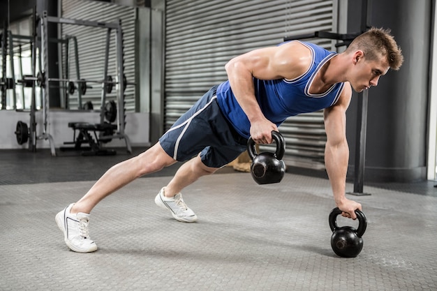 Homem musculoso fazendo push-up com kettlebells no ginásio crossfit