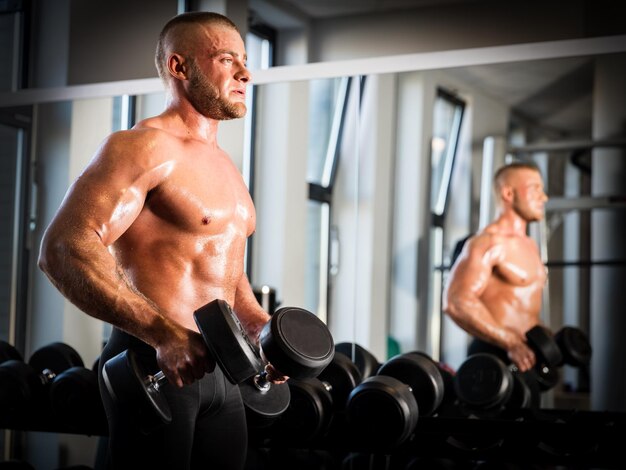 Homem musculoso forte trabalhando com halteres ao lado de um espelho Treinamento de braços e levantamento de pesos em um ginásio Conceito de esporte e musculação