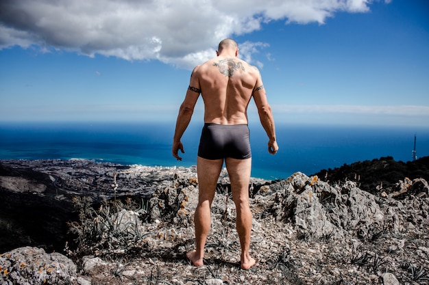 Homem musculoso no topo da montanha com vista para uma cidade e o mar