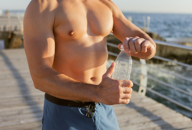 Homem musculoso segurando uma garrafa de água ao nascer do sol na praia