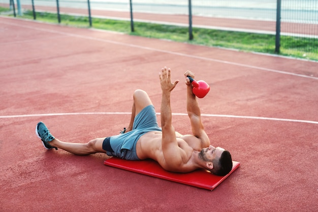 Homem musculoso sem camisa deitado no tapete na quadra e fazendo exercícios com kettlebell pela manhã. conceito de aptidão ao ar livre.