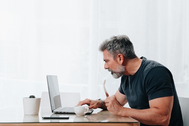 Homem na casa dos 50 anos trabalhando em casa enquanto toma um café e está em videochamada no computador