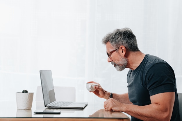 Homem na casa dos 50 anos trabalhando em casa enquanto toma um café e está em videochamada no computador