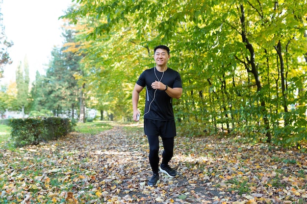 Homem na corrida matinal no parque de outono esportista asiático em roupas esportivas com fones de ouvido e telefone na floresta