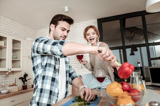 Homem na cozinha. Homem bonito de cabelos escuros e camisa quadriculada se divertindo com vegetais