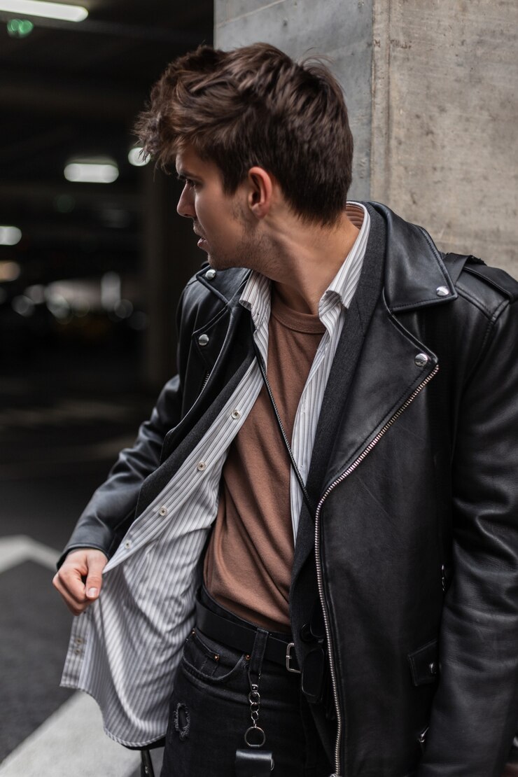 Foto homem na moda moderno jovem hippie com um penteado elegante na jaqueta de couro superdimensionada da moda em uma camisa parece longe, perto de uma coluna vintage na cidade. modelo de cara urbano ao ar livre. moda americana.