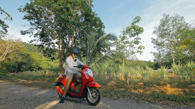 Homem na moto vermelha em roupas brancas dirige na viagem de trilha da floresta. Um turista caucasiano de homens vai de scooter, palmeira tropical próxima. Turismo de passeio de Ásia Tailândia. Aluguel de motocicleta Capacete de segurança.