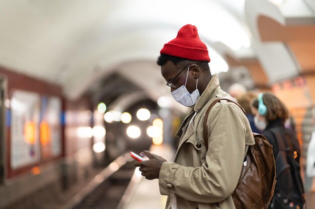 Homem negro à espera de trem na estação de metrô usando telefone móvel usa máscara facial pandemia de covid-19
